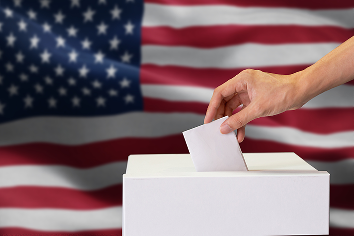 Photo of a hand, placing a piece of paper inside of a ballot box with an American flag in the background