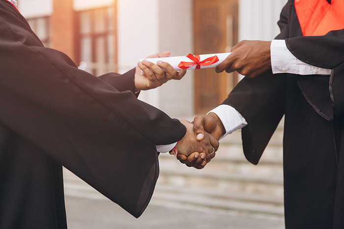 Photo of two graduates in academic gowns exchanging a diploma with a handshake