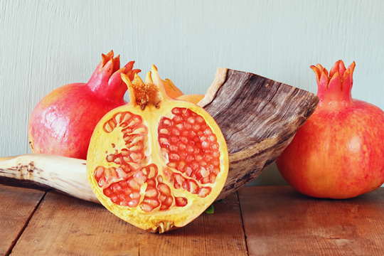 an image of a shofar with two full pomegranates on either side and a pomegranate sliced in half in front of it, all sitting on a table