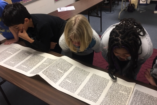 Three children reading a megillah placed on a brown table 