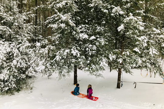 Photo of two kids sitting on a snow board surrounded by a lot of snow and trees